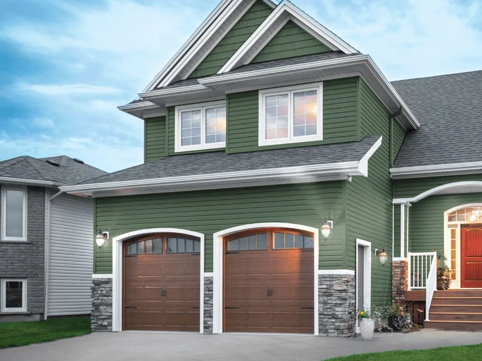 A modern two-story house with Shamrock-colored siding, featuring a double garage with brown doors, stone accents, and a red front door with a small porch.