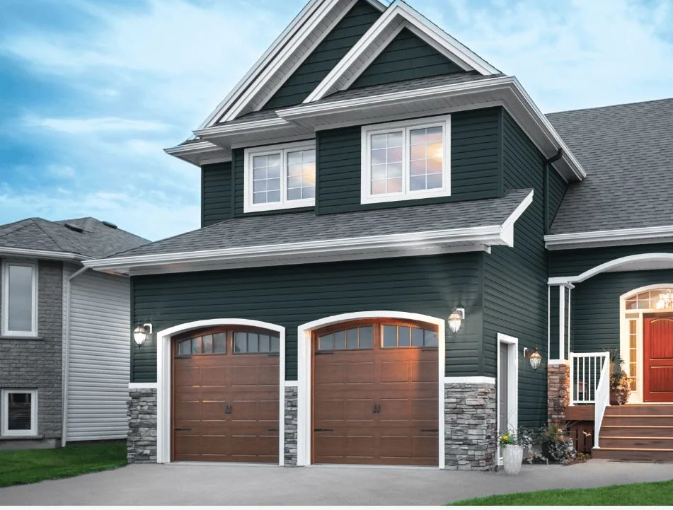 A modern two-story house with wedgewood-colored siding, featuring a double garage with brown doors, stone accents, and a red front door with a small porch.