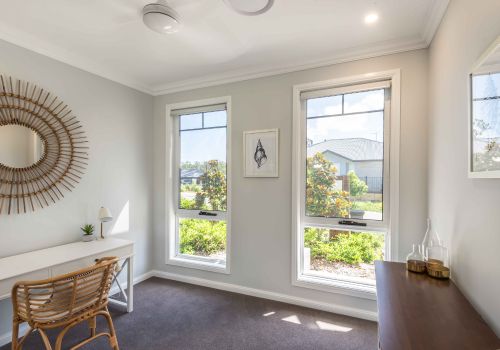Awning windows in a bright home office with light walls, a wicker chair, wooden desk, and decorative mirror, overlooking a green outdoor view.