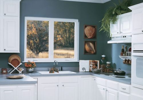 Casement window in a modern kitchen with white cabinets, blue walls, and a scenic autumn view, featuring countertop decor and hanging plants.