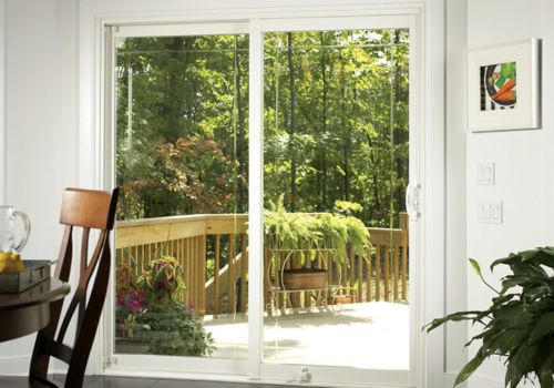 Sliding patio door with large glass panels in a modern room, opening to a wooden deck with plants and a scenic forest view.