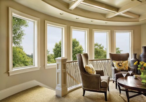 Picture windows in a cozy living room with a curved wall, featuring a large armchair, round table, and scenic view of trees outside.