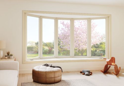 Bay and bow window with five panels in a bright living room, overlooking a garden with blooming trees, featuring a tufted ottoman and rocking horse.