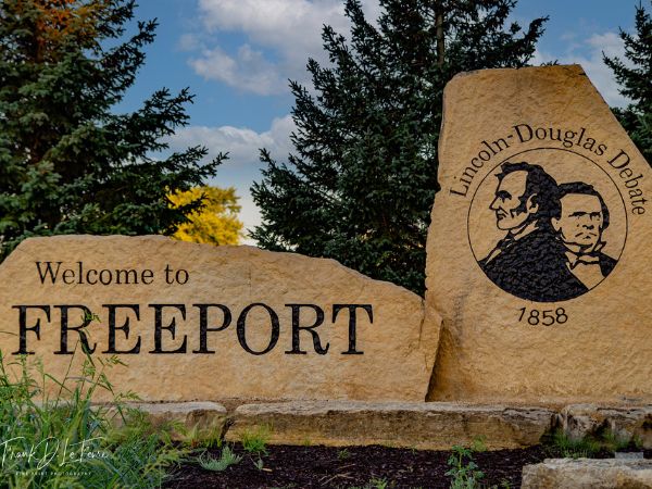 Welcome to Freeport stone sign with Lincoln-Douglas Debate emblem and wooded background for roofing services